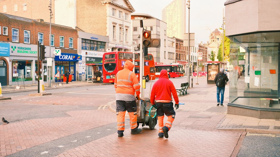 A male street cleaner wearing a high-visibility orange jacket and trousers, holding a long-handled broom, walking across a city road near the entrance of a modern glass building. The building features
